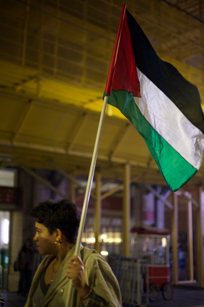 A person holds a Palestinian flag against a backdrop of a softly lit urban environment at night.