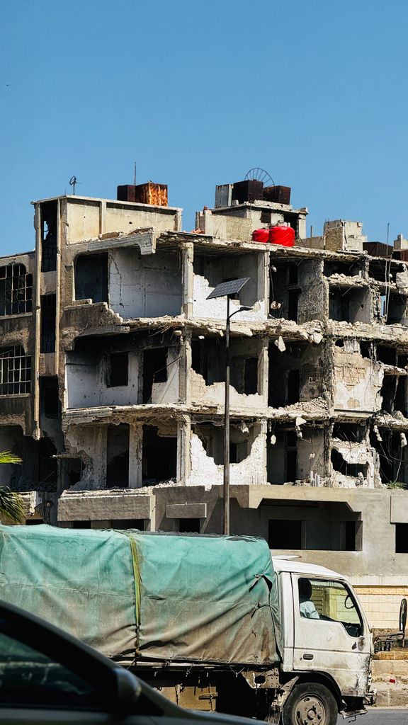 Desolate war-damaged building in Homs, Syria, with a truck in foreground.