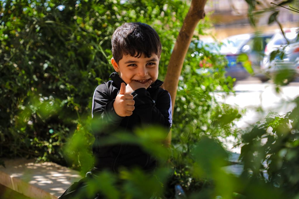 Young boy smiling and giving thumbs up outdoors amidst lush green foliage.